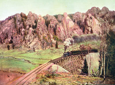 In a photo that was long ago tinted for effect, a Short Line 
to Cripple Creek train chugs over one of the lines 31 timber 
railroad bridges as it passes through Cathedral Park in 1904. The Short Line, built by Irving Howbert as part of a
fierce railroad competition for Cripple Creek gold, evolved into Gold Camp Road.